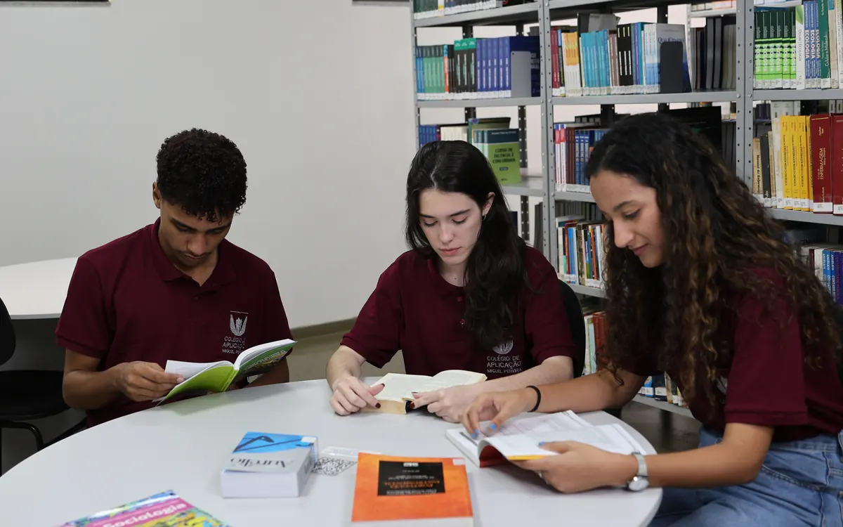 Três alunos de uniforme estudam juntos na biblioteca