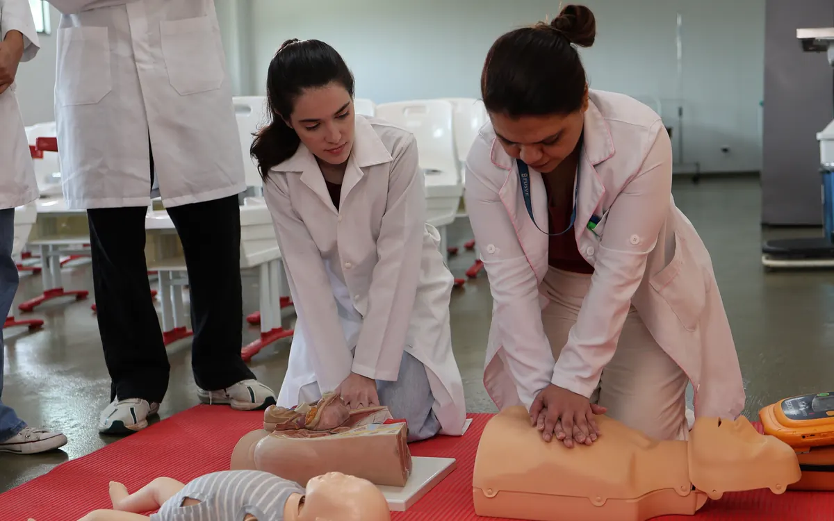 Alunas de jaleco praticam massagem cardíaca em bonecos de treinamento no laboratório de saúde
